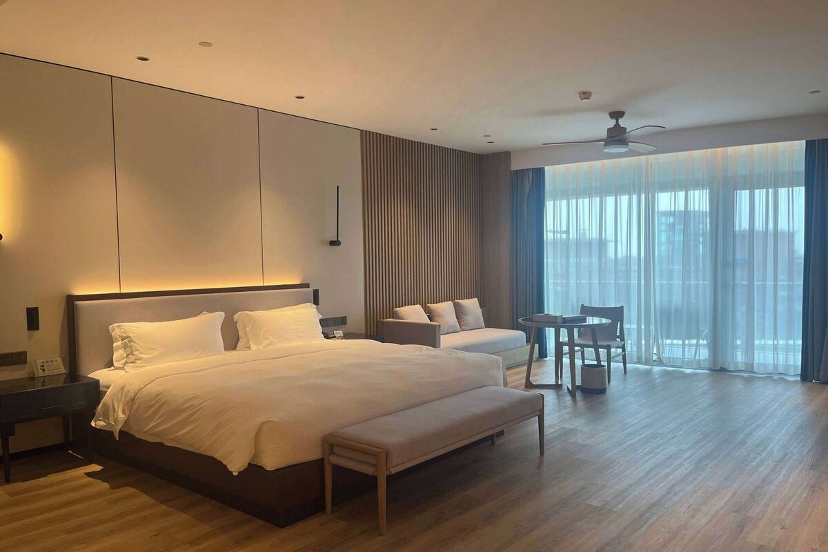 Ein modernes Hotelzimmer mit einem großen Bett, einem Nachttisch, einem Schreibtisch, einem Sessel, einem kleinen Tisch und einem großen Fenster mit Vorhängen.
