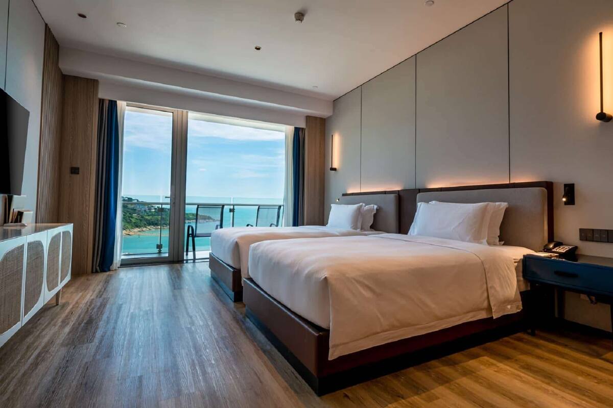 Ein modernes Hotelzimmer mit einem großen Bett, einem Schreibtisch und Blick auf den Ozean durch eine Schiebetür.