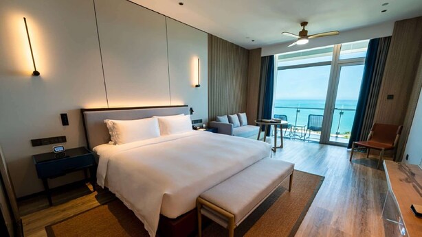 Une chambre d’hôtel moderne dotée d’un grand lit, de tables de chevet, d’un bureau, d’un canapé, d’un balcon avec vue sur la mer et d’un ventilateur de plafond.