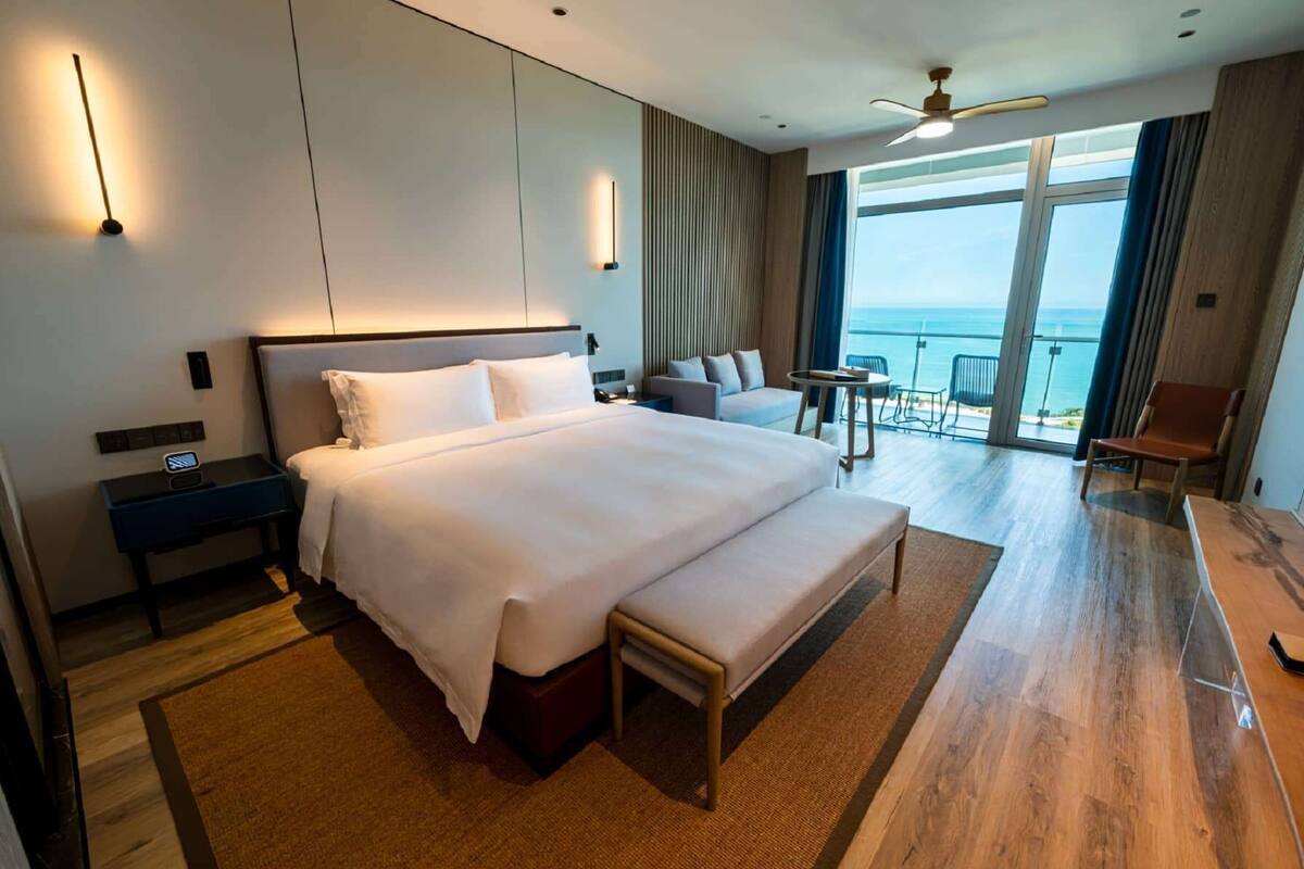 Ein modernes Hotelzimmer mit einem großen Bett, Nachttischen, einem Schreibtisch, einer Couch, einem Balkon mit Meerblick und einem Deckenventilator.