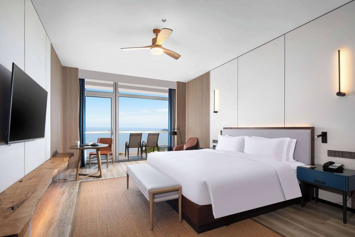 Ein modernes Hotelzimmer mit einem großen Bett, einem Schreibtisch, einem Sessel, einem Fernseher und Meerblick.