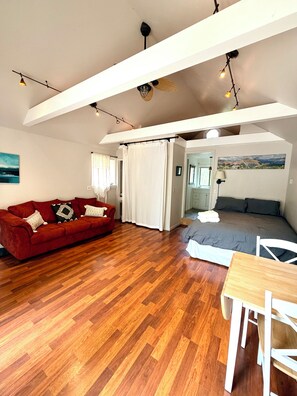 Una stanza con un divano rosso, un tavolo da pranzo in legno, un letto con materasso grigio e una tenda bianca.