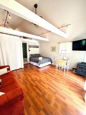 A spacious room with a wooden floor, a bed, a red sofa, a dining table with chairs, and a television. - The Guest Cottage in Lake City (Lake City)