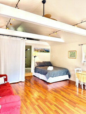 Une chambre à coucher avec un lit, une chaise rouge, un plancher en bois, une table blanche et un tableau accroché au mur.