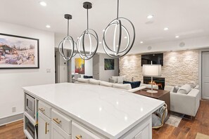 A modern kitchen with a large island, pendant lighting, a stone accent wall, a fireplace, and a living area with a sofa and a painting. - BRIGHT, Renovated, In the Heart of Aspen Walkable (Aspen)