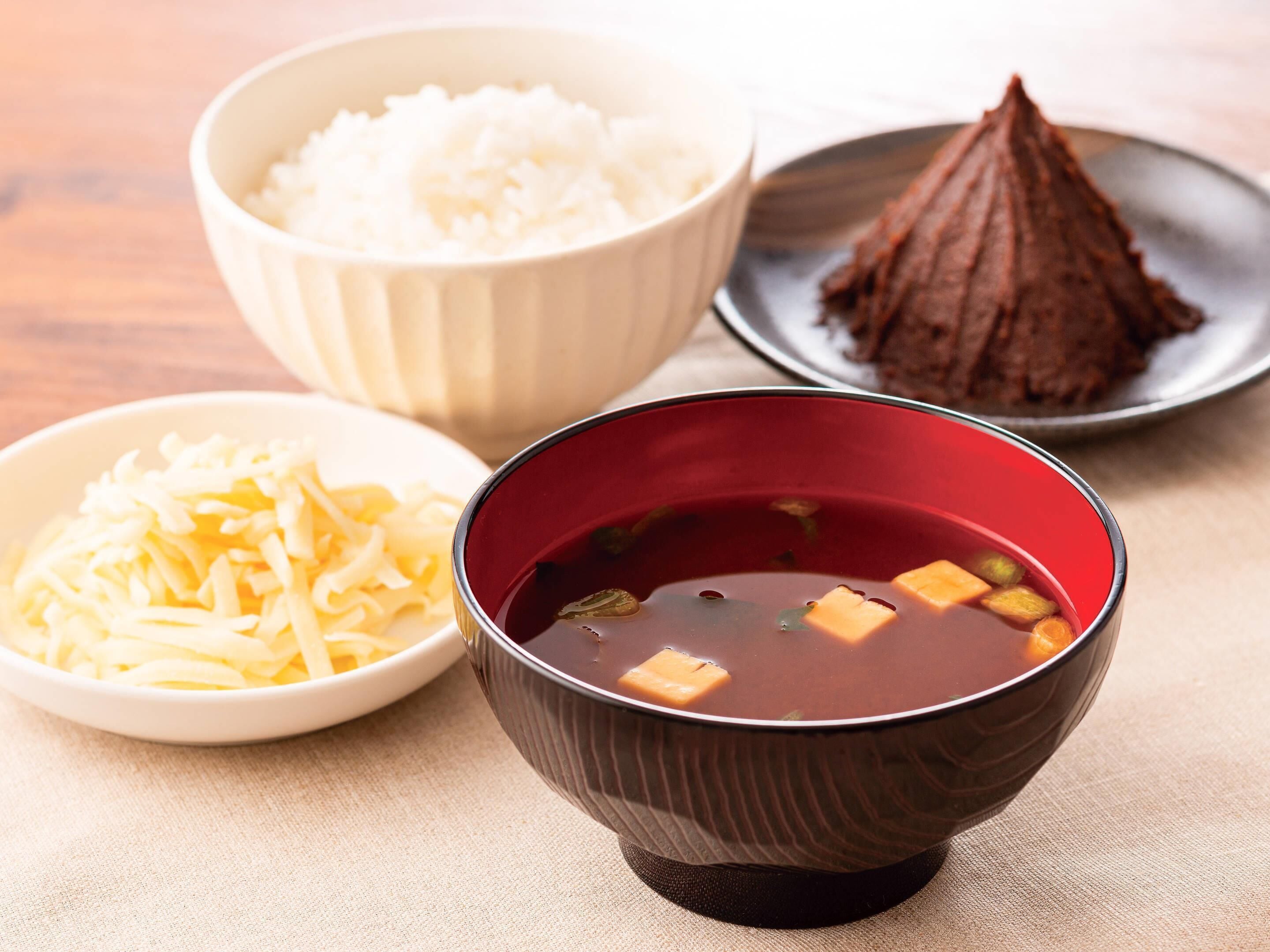 Une table avec trois bols contenant différents aliments : un bol de riz, un bol de soupe foncée et un bol contenant un aliment de forme triangulaire.