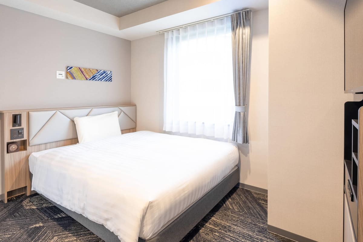 A hotel room with a bed, a bedside table, a clock, a power outlet, a window with curtains, and a wall-mounted TV.