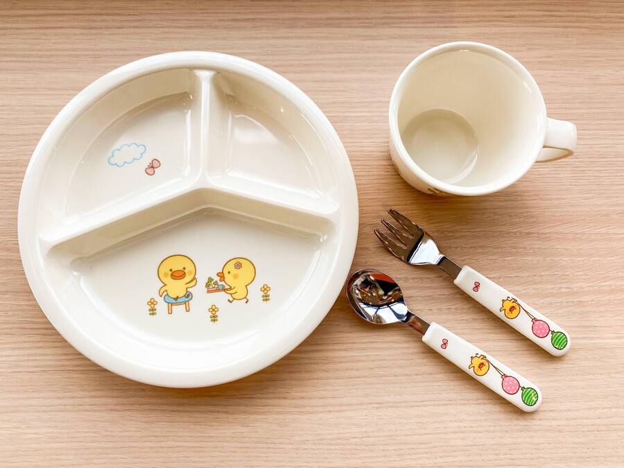 A white plate with a divided section and two cartoon ducks, a white cup, and a fork with a handle decorated with cartoon ducks.