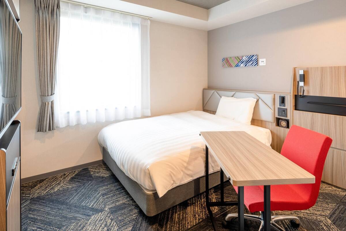 A hotel room with a bed, a desk, a chair, a TV, and a window with curtains.
