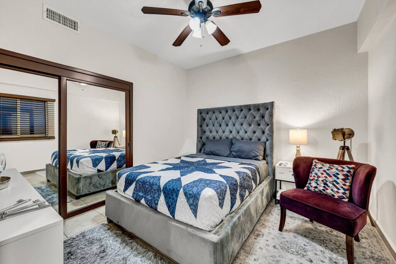 A bedroom with a large bed, a ceiling fan, a mirror, a desk, and a chair.