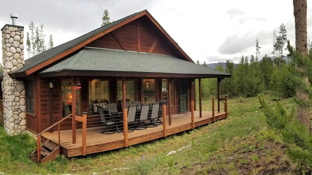 Une cabane en rondins avec une cheminée en pierre, une véranda couverte avec des sièges et un paysage montagneux en arrière-plan.