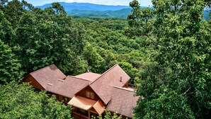 A lodge with a prominent wooden structure surrounded by dense forest, featuring a large deck and a staircase leading to the entrance. - Game Rooms + Views: Spacious Mtn Cabin in Murphy! (Murphy)