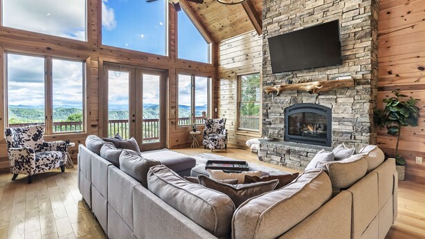 A spacious living room with a large sectional sofa, a stone fireplace, and a flat-screen TV mounted on the wall. - Game Rooms + Views: Spacious Mtn Cabin in Murphy! (Murphy)