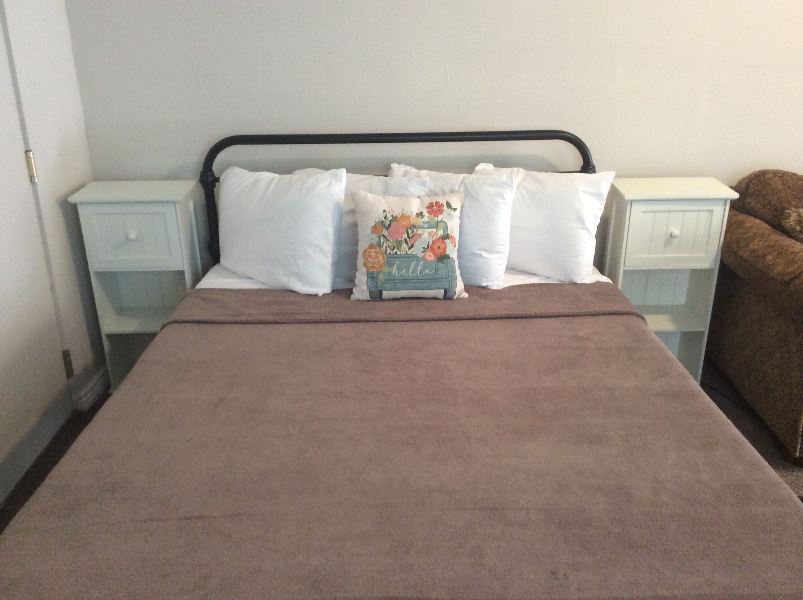 A bed with a brown blanket, white pillows, and a floral pillow. There are two bedside tables with white drawers.