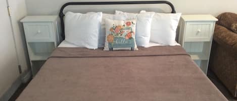 A bed with a brown blanket, white pillows, and a floral pillow. There are two bedside tables with white drawers.