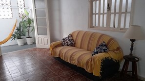 Uma sala de estar com sofá, duas almofadas, luminária, janela com persianas, porta e plantas em vasos.