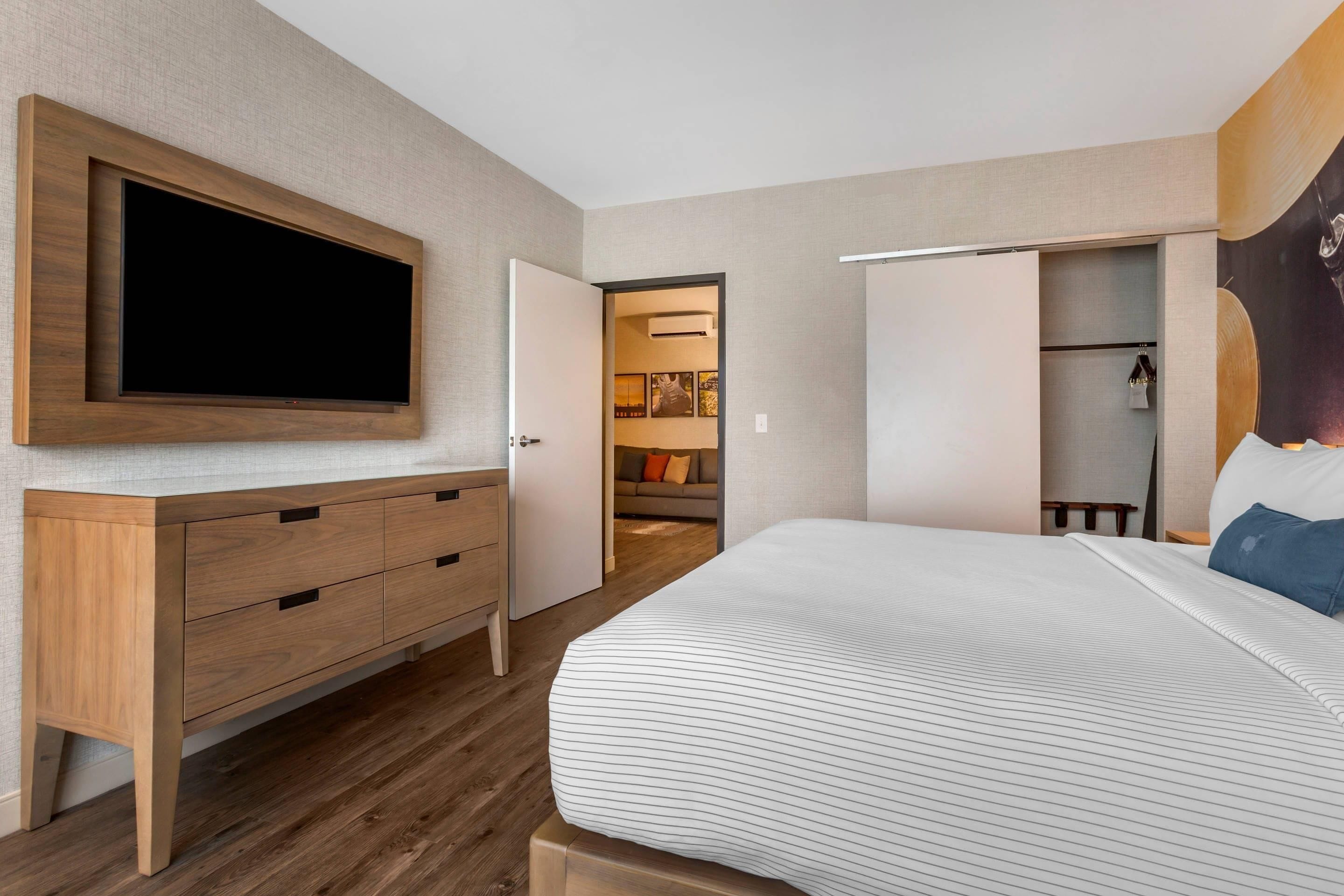 Un dormitorio con una televisión de pantalla plana grande montada en la pared, un aparador de madera, una cama con ropa de cama a rayas y un vestidor con espejo y ropa colgada.