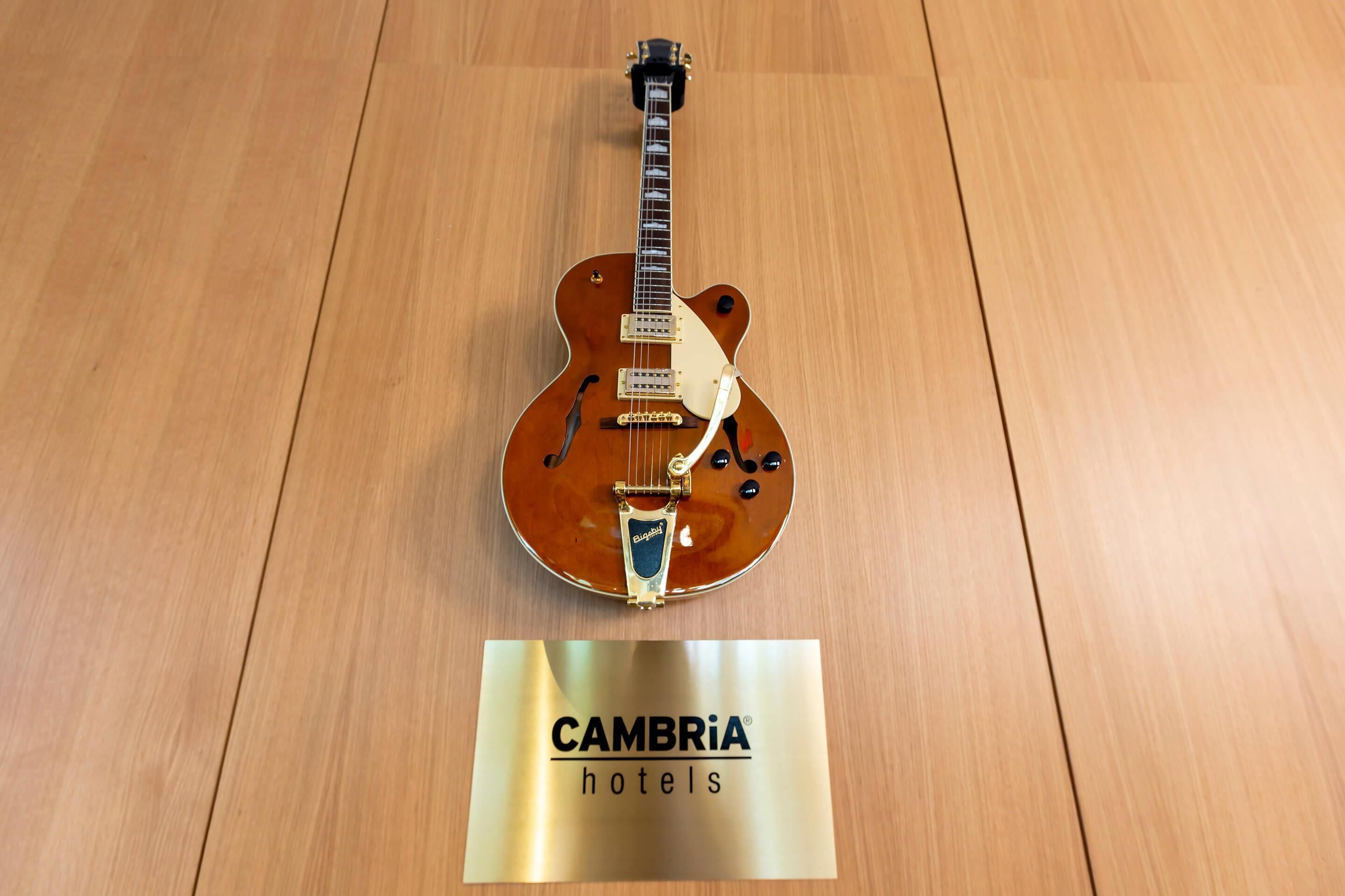 Une guitare posée sur un plancher en bois, avec une pancarte indiquant « Hôtels CAMBRIA ».