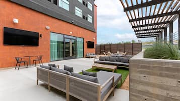 Un patio exterior con mobiliario moderno, un televisor de pantalla plana montado en el edificio y una zona de estar cubierta.