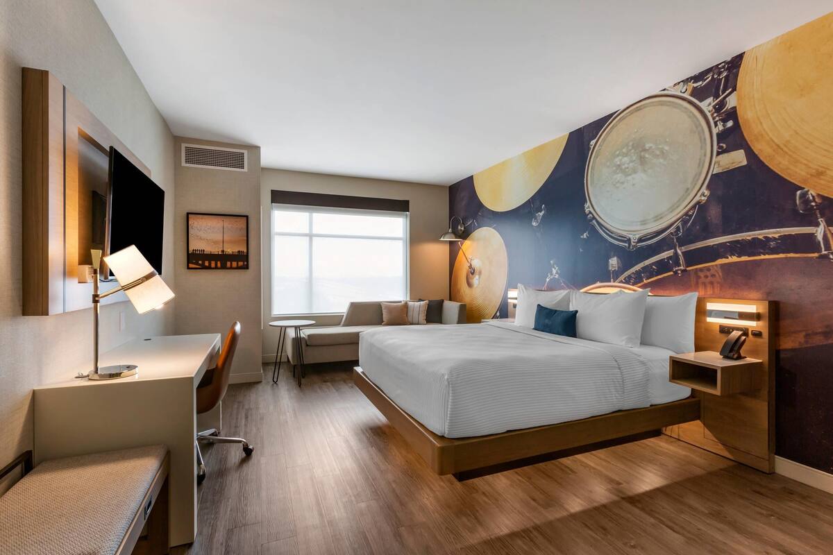 A modern hotel room with a bed, desk, and chair, a large window, and a wall mural.