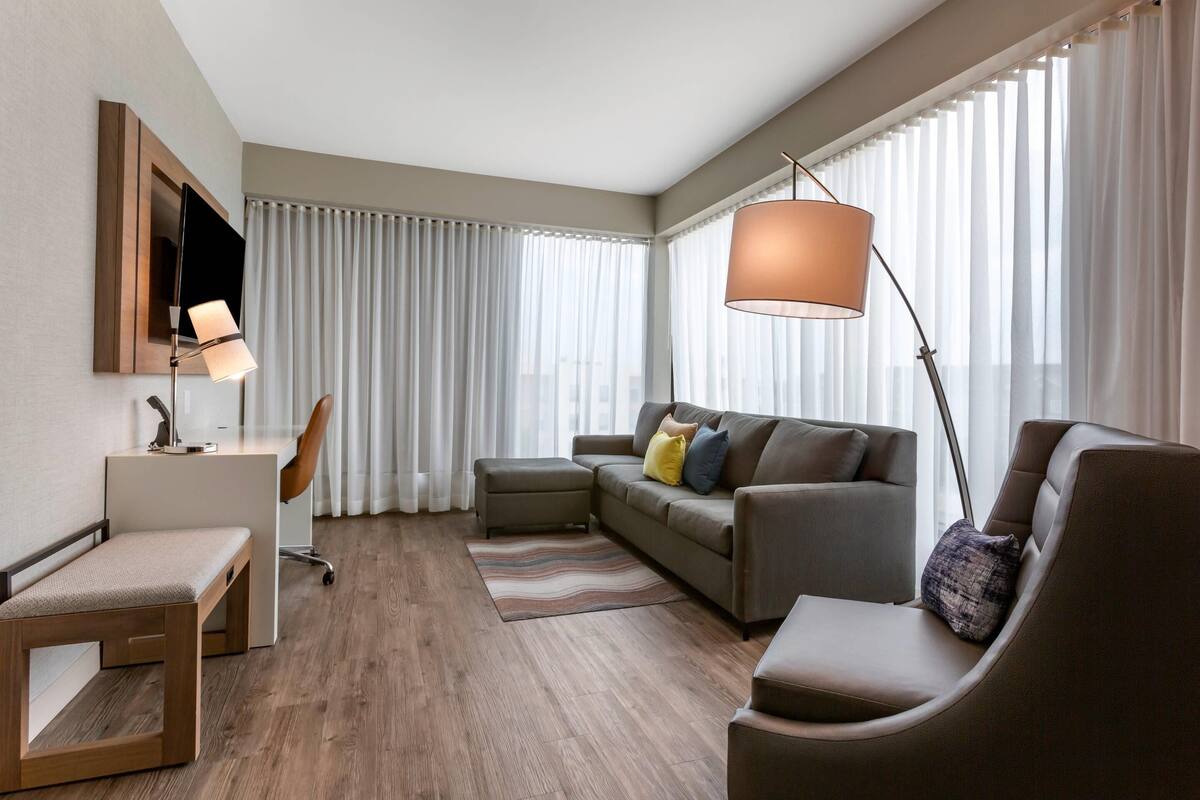 A modern hotel room with a sofa, armchair, desk, and a large window with curtains.