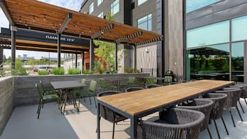 Un patio al aire libre con una mesa de madera y sillas de mimbre, un espacio de estar cubierto y vistas al exterior del edificio.
