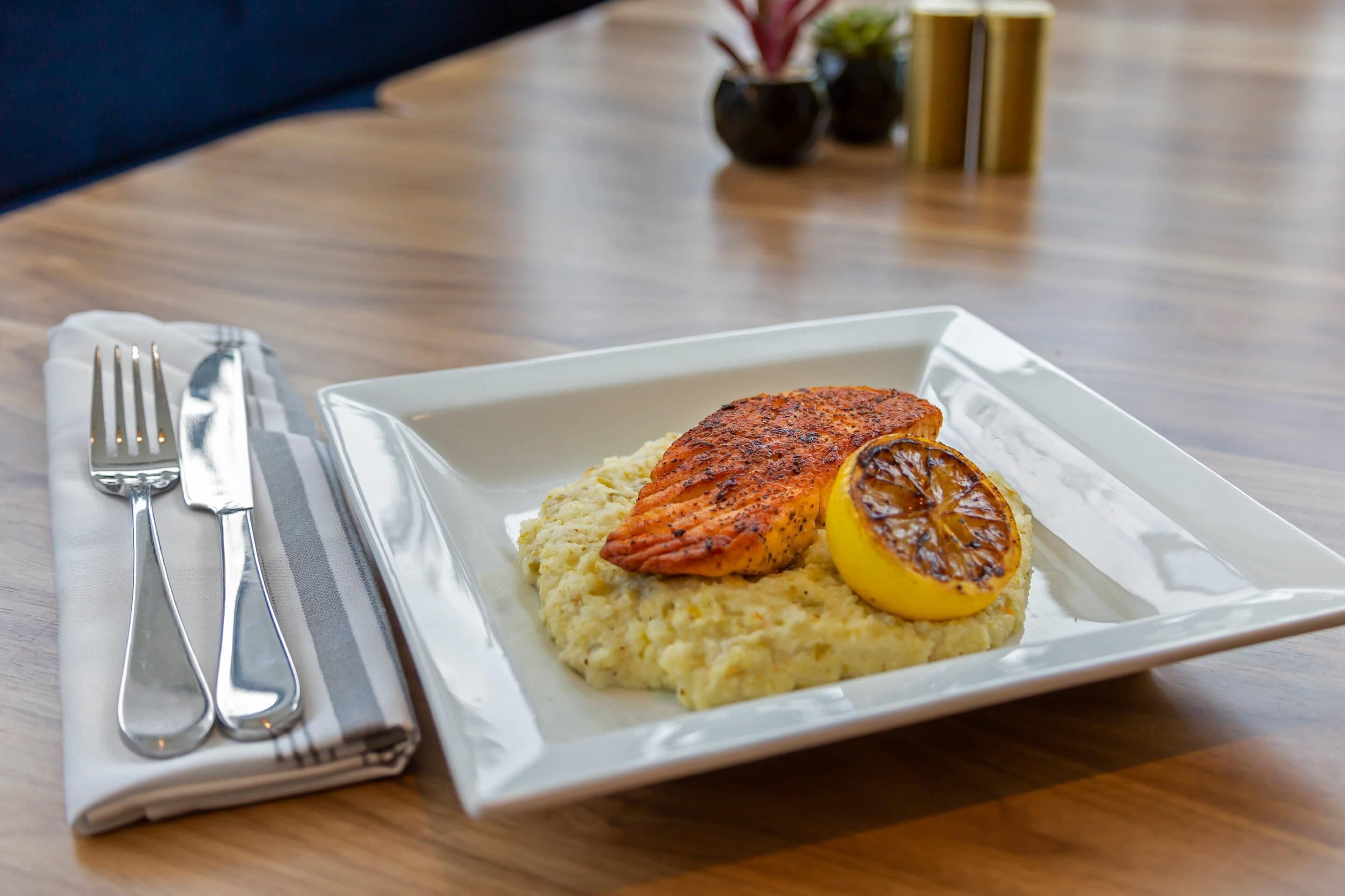 A white plate with salmon and a lemon slice on a bed of mashed potatoes, placed on a wooden table.