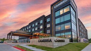 Un hotel moderno con un llamativo letrero de "CAMBRIA", un acceso cubierto y un césped bien cuidado.
