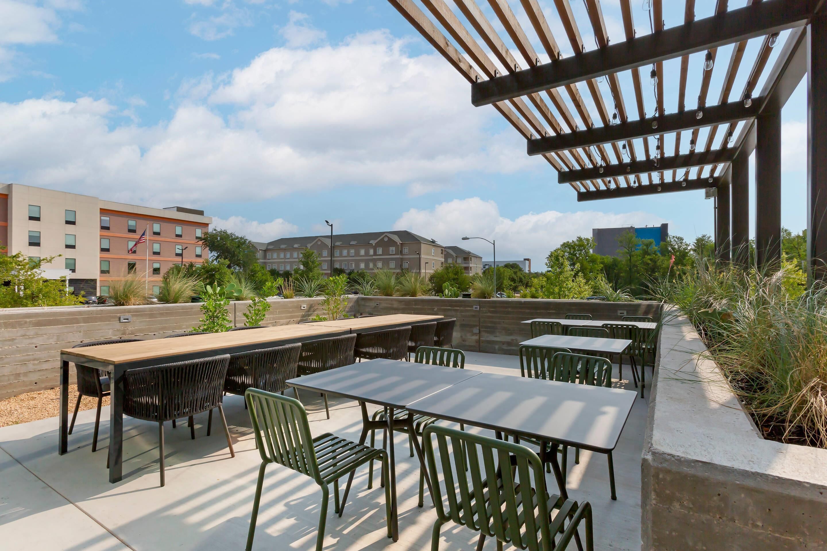 An outdoor patio area with tables and chairs, a pergola structure, and a view of buildings in the background.