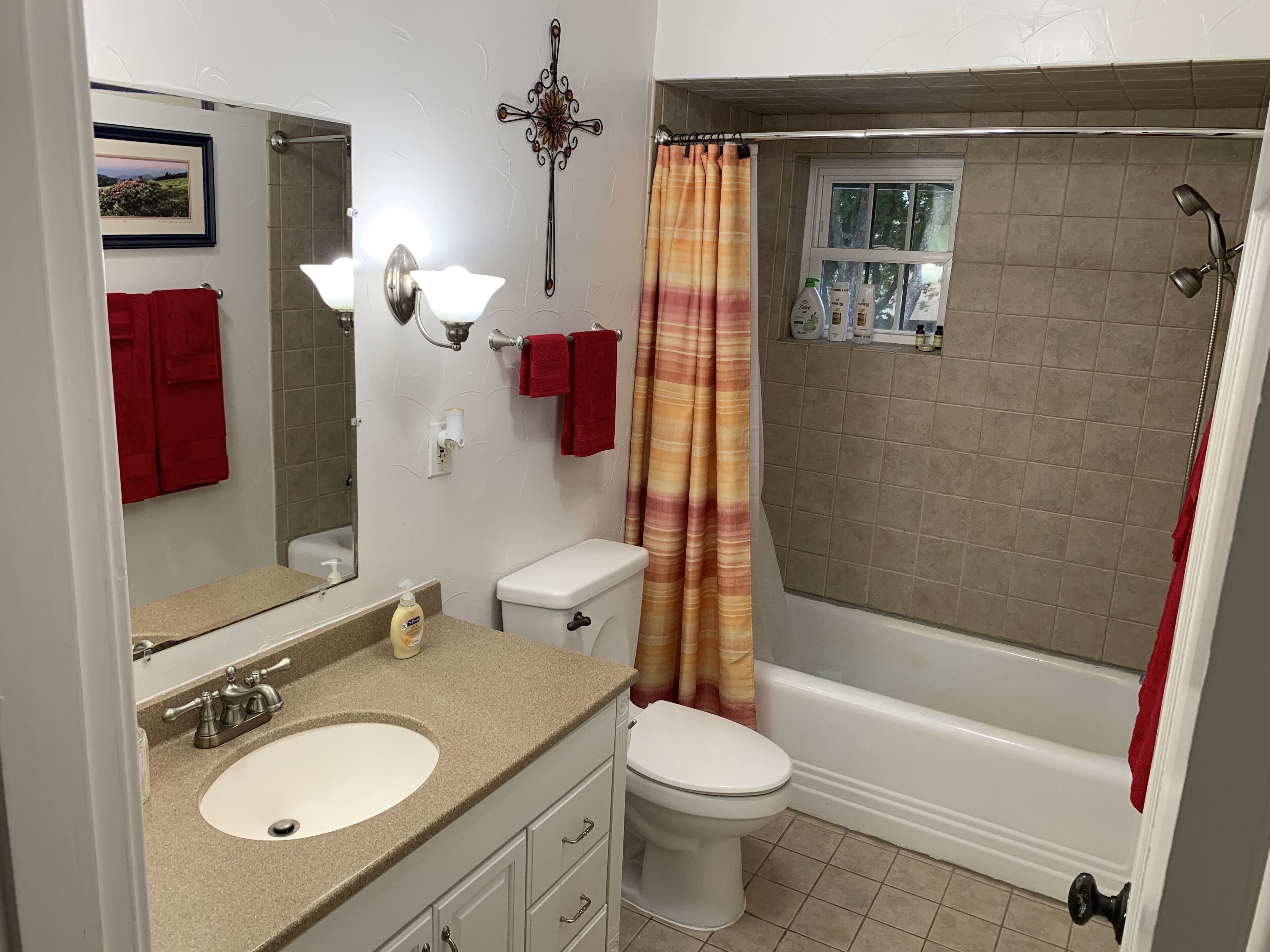 Une salle de bain avec un lavabo, des toilettes et une douche. Une serviette rouge est accrochée au mur, il y a un tableau encadré et un rideau de douche.