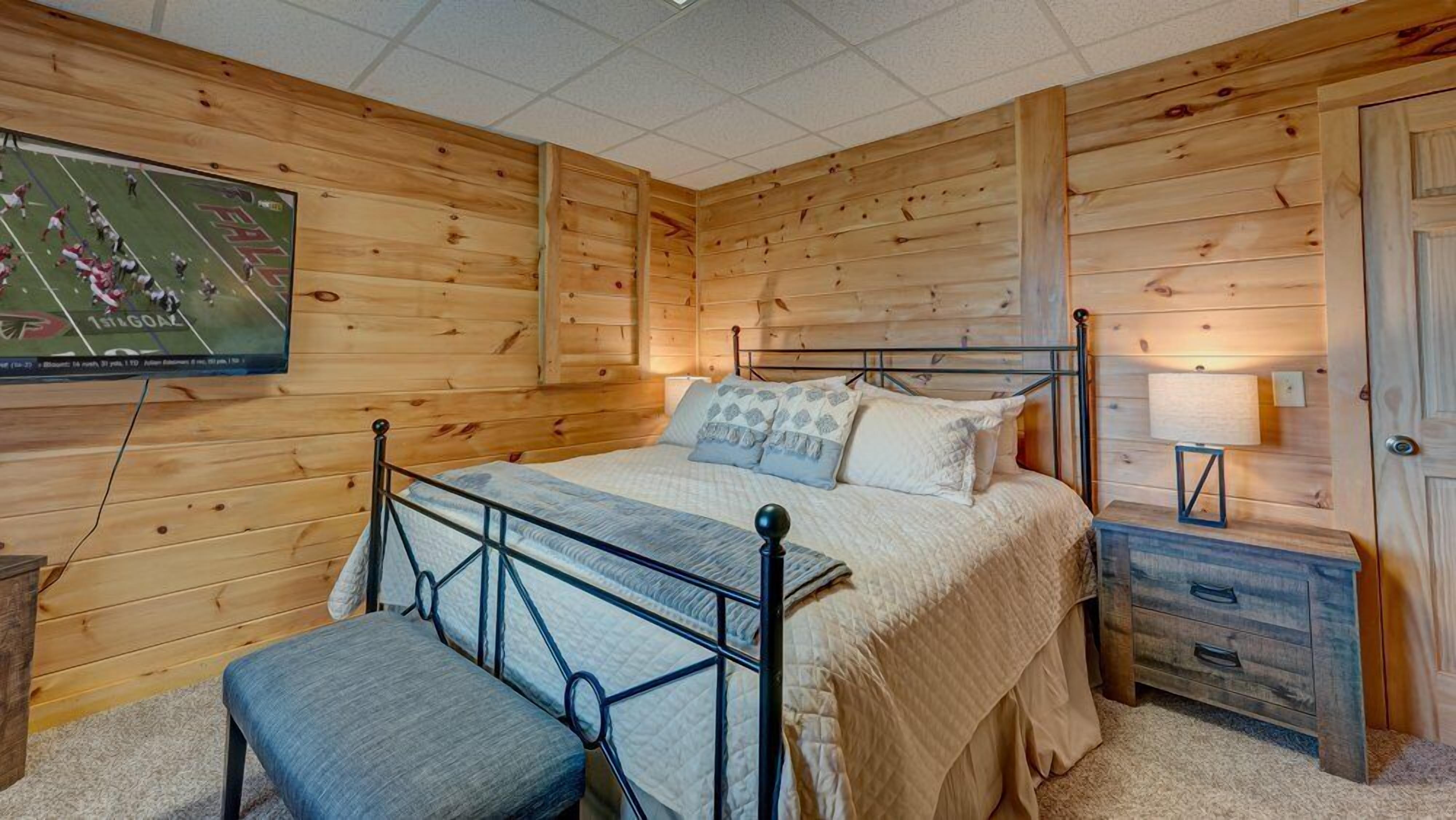 A wooden-paneled room with a bed, a bench, a nightstand, and a TV on the wall.