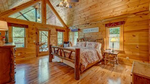 Ein Schlafzimmer in einer Blockhütte mit einem Bett, einem Nachttisch, einer Lampe und einem Fenster mit Blick auf Bäume.