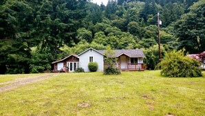 A two-story house with a front porch, surrounded by a grassy yard and dense forest.