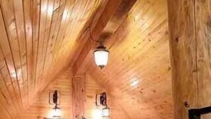 A cozy attic bedroom with wooden walls and a sloped ceiling. There is a bed with a gray bedspread and two pillows. Two wall-mounted lamps provide lighting.