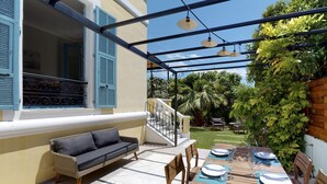 Een overdekt terras met een eettafel voor vier personen, een bank en een trap naar een balkon.