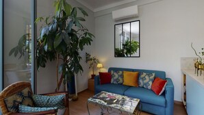 Een woonkamer met een blauwe bank, een rieten stoel, een glazen salontafel en een grote plant.
