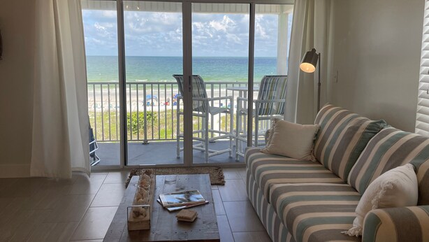 A room with a large sliding glass door leading to a balcony with a view of the ocean. There is a striped sofa and a wooden coffee table with books and decorative items.