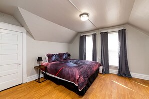 Cozy loft-style bedroom with a bed, bedside table, and lamp, featuring a large window with curtains.