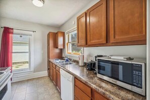 A kitchen with wooden cabinets, a granite countertop, a microwave, a dishwasher, and a window with a red curtain. - Entire Home I 3 Bedroom I 2 Bath (Lansing)