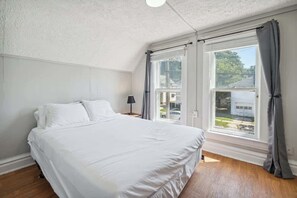 A bedroom with a bed, a nightstand, a lamp, and a window with curtains. - Entire Home I 3 Bedroom I 2 Bath (Lansing)