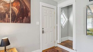 Een gang met een witte deur, een houten vloer, een deurmat en een ingelijst schilderij aan de muur.