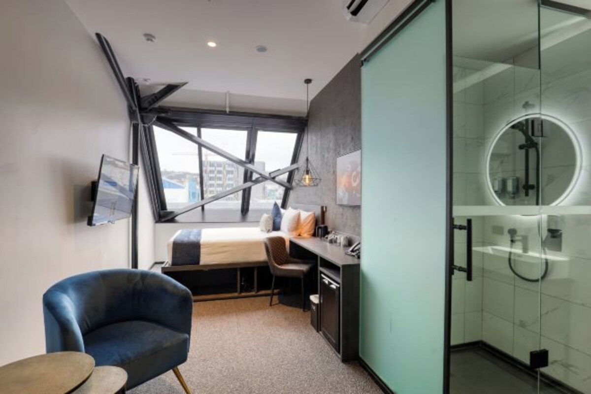 A modern hotel room with a bed, desk, chair, and a glass-enclosed shower area.