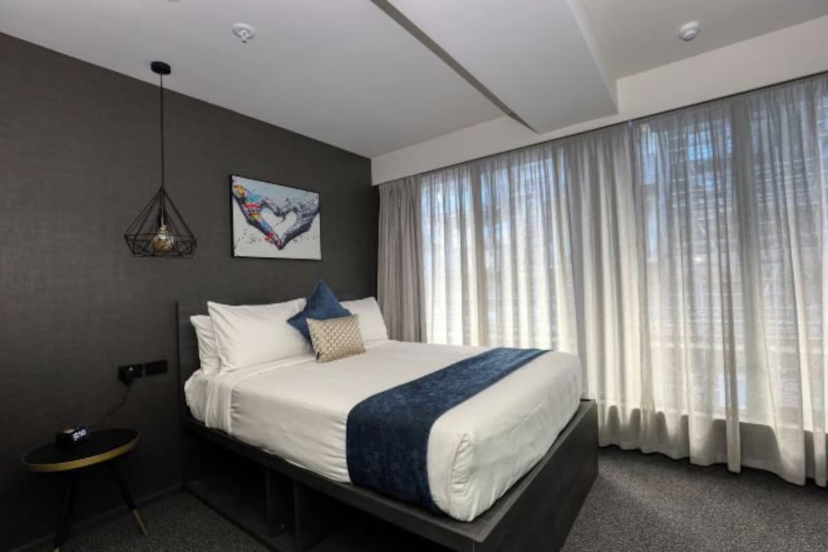 A modern hotel room with a large bed, a bedside table, a wall-mounted light fixture, and a window with sheer curtains.