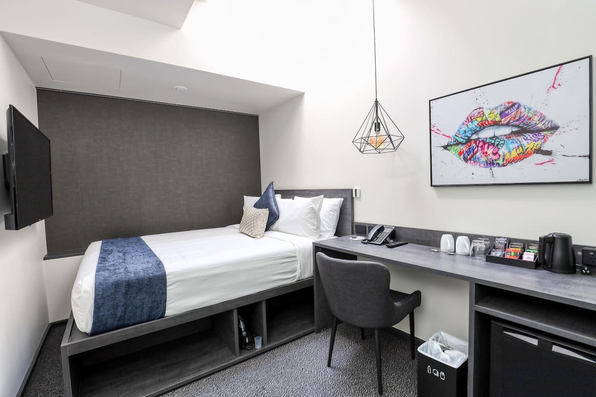 Moderni hotellihuone, jossa on sänky, työpöytä, tuoli ja suuri huuliaiheinen taideteos seinällä.