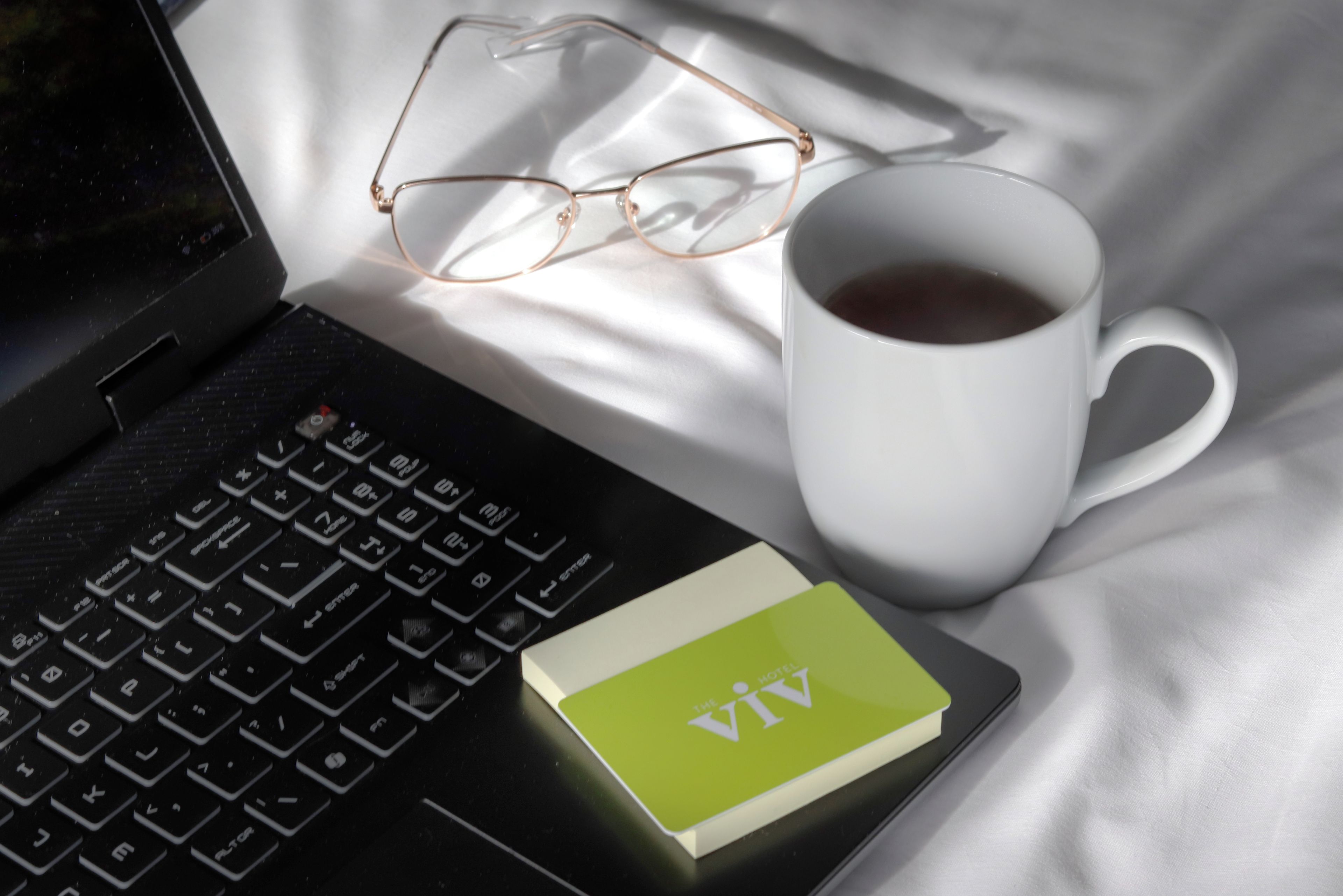 Un ordenador portátil, un par de gafas, una taza blanca y una nota adhesiva sobre una cama.