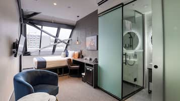 A modern hotel room with a bed, desk, chair, and a glass-enclosed shower.