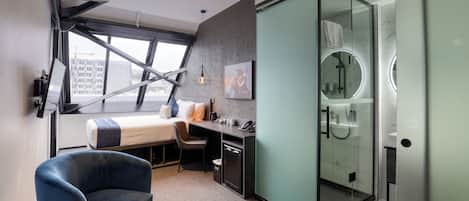 A modern hotel room with a bed, desk, chair, and a glass-enclosed shower.