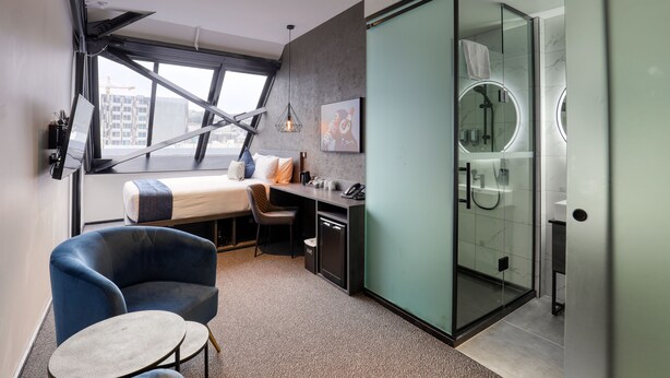 A modern hotel room with a bed, desk, chair, and a glass shower enclosure.