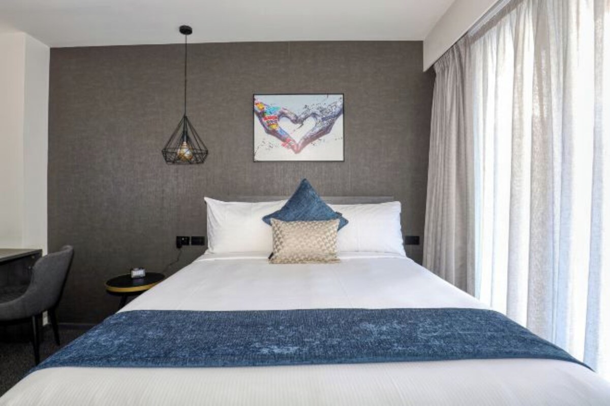 A hotel room with a large bed, a desk, a chair, a small table, a hanging light fixture, and a heart-shaped artwork on the wall.