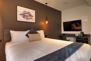 Ett modernt hotellrum med en stor säng, en platt-TV, ett skrivbord med en kaffebryggare och ett unikt konstverk på väggen.